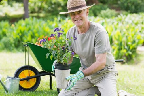 A middle-aged man wearing a wide-brimmed straw hat, light grey t-shirt, and gardening gloves is kneeling on a well-maintained lawn in a backyard garden, holding a potted flowering plant with purple and red blooms. Behind him, there is a garden wheelbarrow with a bright yellow wheel, filled with more container plants, and a metal watering can resting on the grass. The garden features a lush, green lawn bordered by correctly trimmed hedges or shrubs, with tall trees and dense foliage in the background providing shade and a natural setting. The scene suggests outdoor gardening activity on a clear day with bright, natural lighting, emphasizing a neat and cared-for outdoor space. This setup visually supports gardening services offered by Gardening Services Sydenham, highlighting lawn care, planting, and garden maintenance in a typical residential outdoor environment in Sydenham or nearby postcode areas.